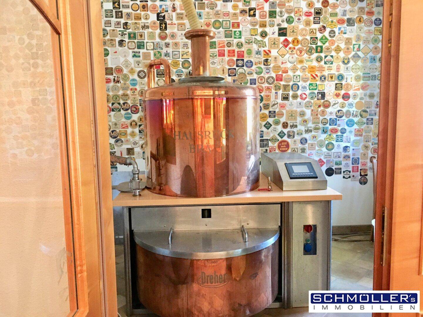 Kupferner Braukessel in einem Raum mit einer Wand voller Bieretiketten, deutet auf eine Brauerei hin.