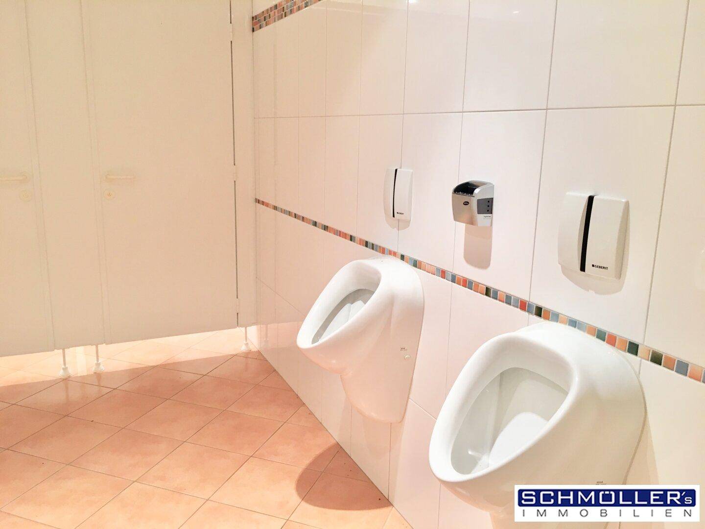 Herren-WC mit zwei Urinalen, weißen Fliesen und farbiger Bordüre.