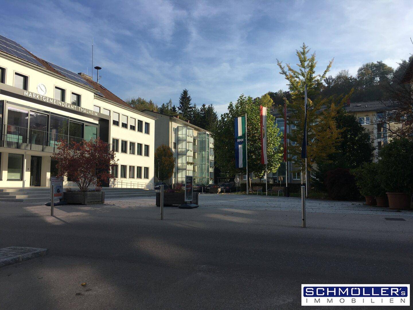 Sonniger Stadtplatz mit modernen Gebäuden, Bäumen und wehenden Flaggen unter blauem Himmel.