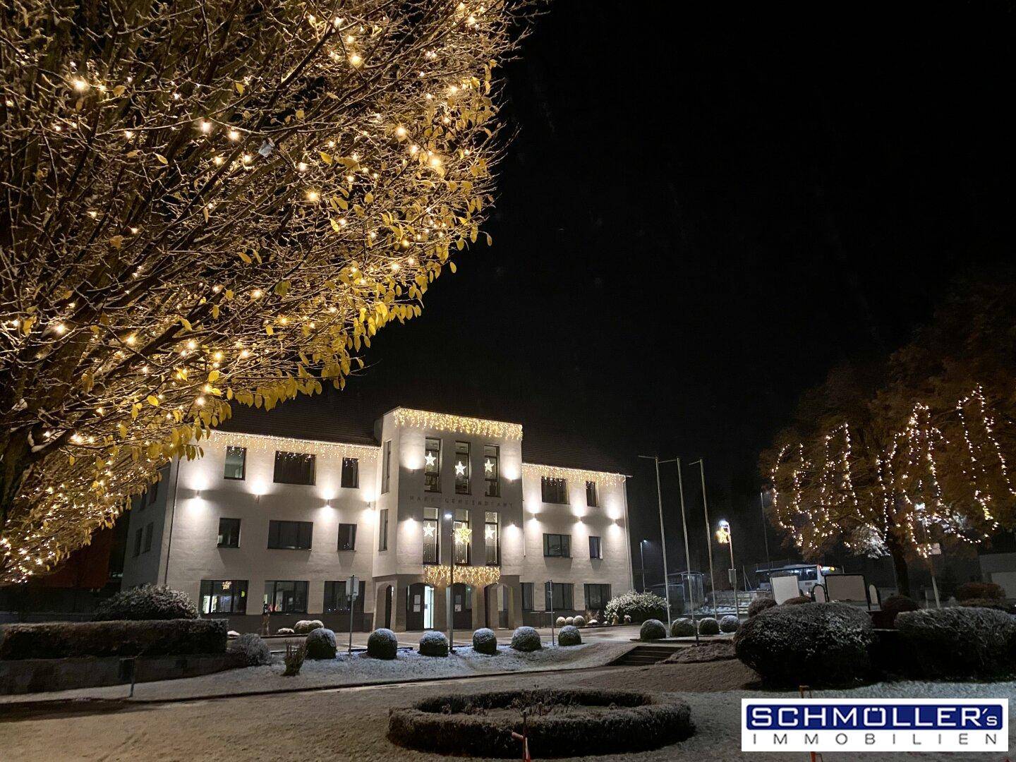 Festlich beleuchtetes Gebäude in einer verschneiten Nacht mit Lichterketten an den Bäumen.