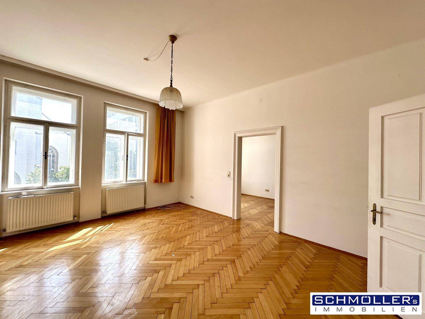 Geräumiges Wohnzimmer mit Fischgrätparkett, hohen Fenstern und viel Tageslicht.
