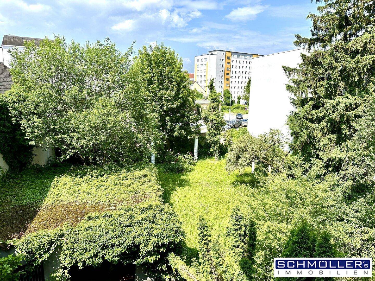 Grüne Gartenfläche mit Bäumen und Sträuchern, umgeben von Wohngebäuden im Hintergrund.