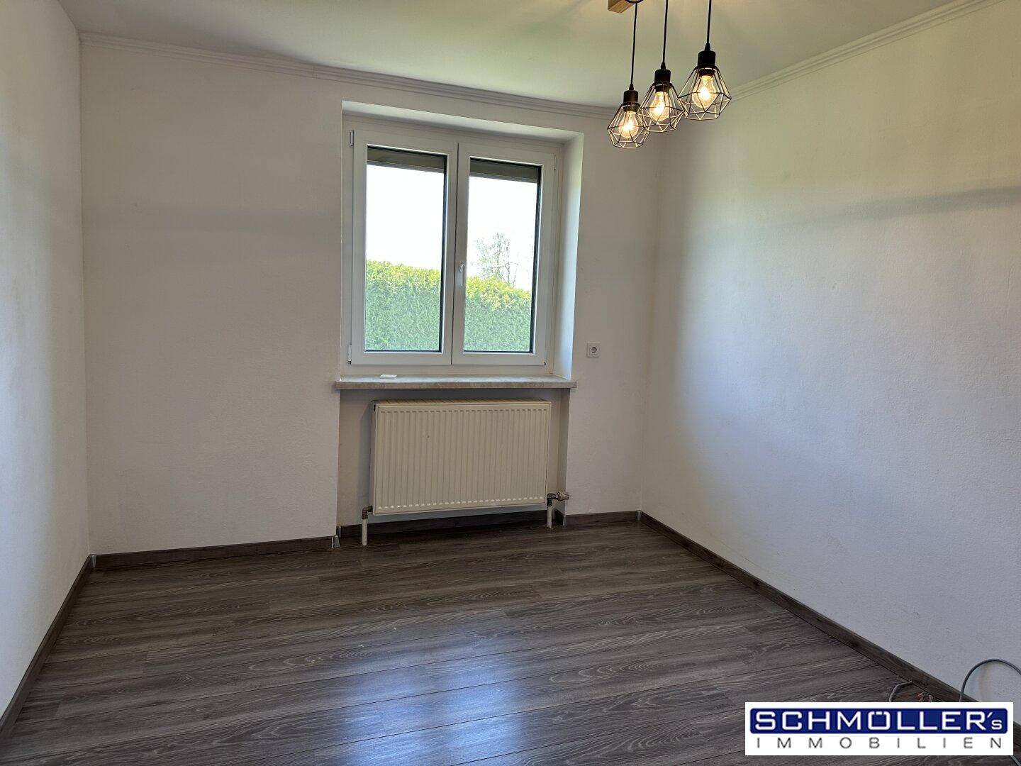 Helles Zimmer mit Laminatboden, weiß gestrichenen Wänden und einem Fenster mit Blick ins Grüne.