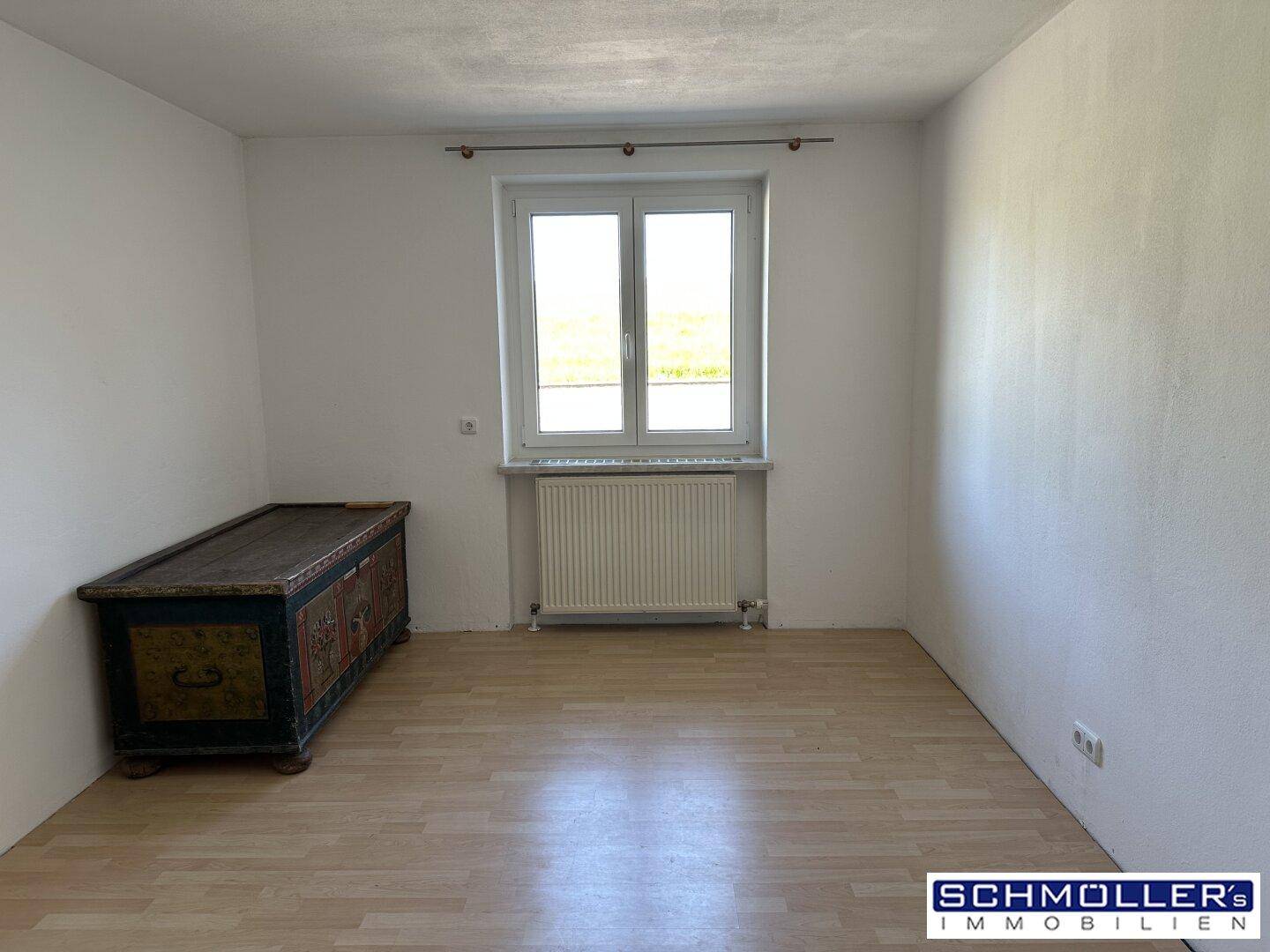 Helles Schlafzimmer mit Parkettboden, weiß gestrichenen Wänden und einem Fenster, das viel Licht hereinlässt.