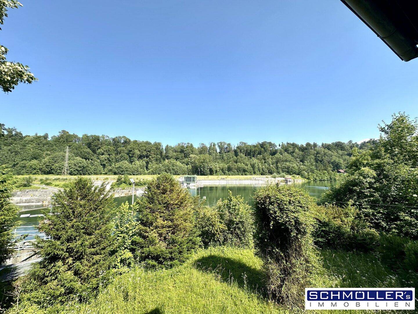 Panoramablick auf einen Stausee und bewaldete Hügel unter klarem blauem Himmel.