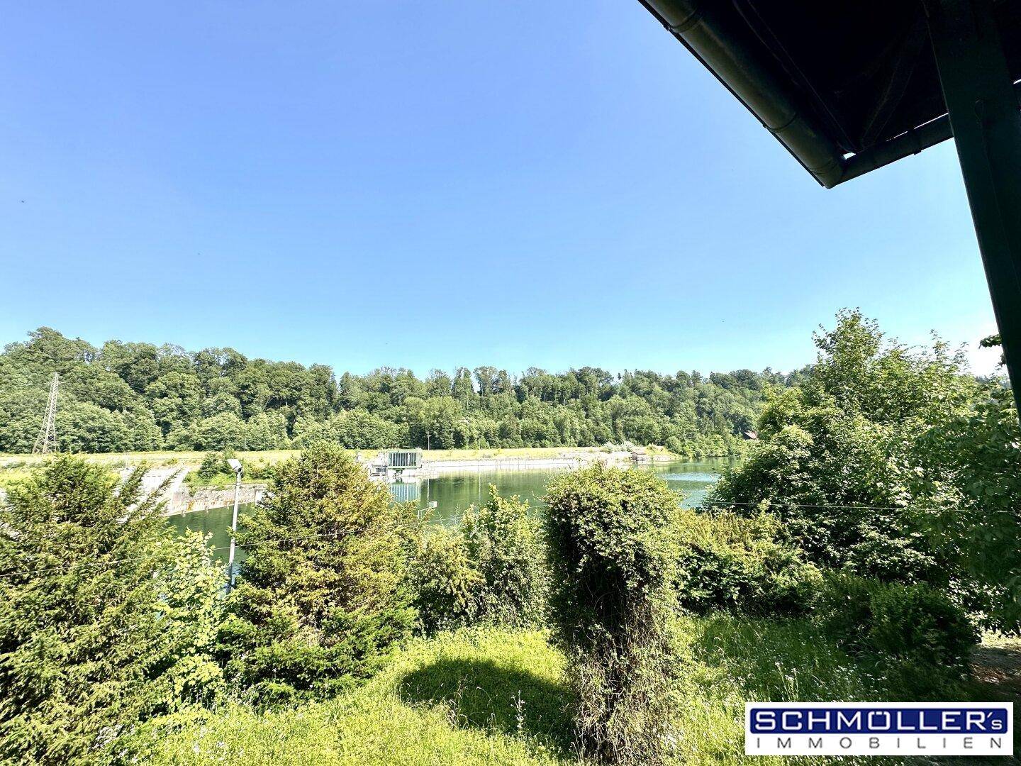 Panoramablick auf einen See oder Fluss mit bewaldeten Ufern und einem Damm im Hintergrund.