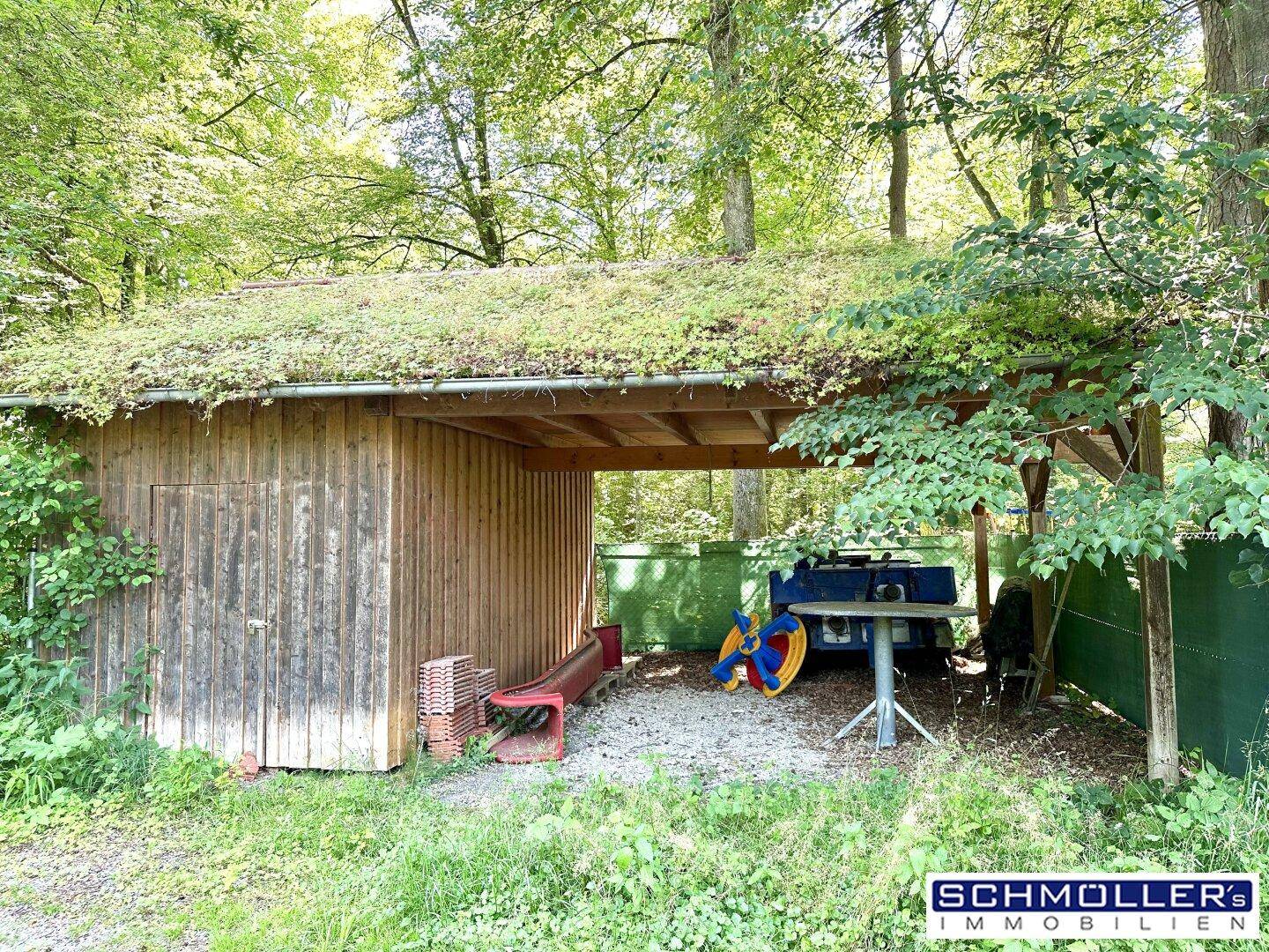 Rustikaler Holzschuppen mit begrüntem Dach und überdachtem Bereich im Grünen, umgeben von Bäumen.