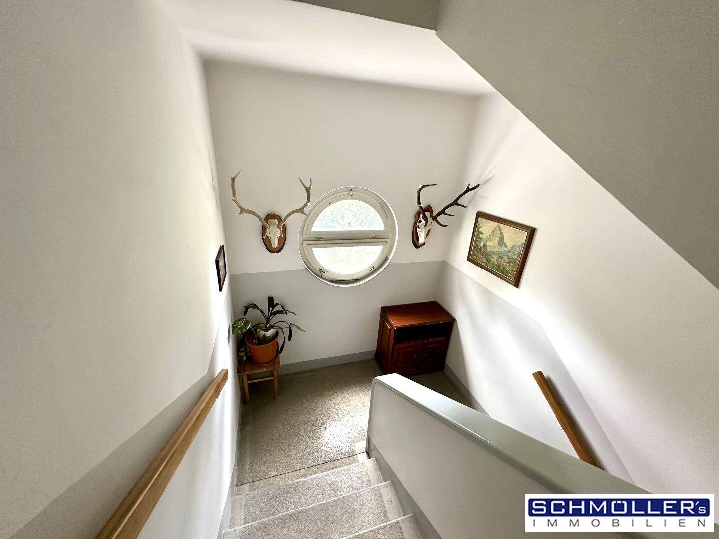 Treppenhaus mit Teppichboden, Rundfenster und Jagdtrophäen als Dekoration.