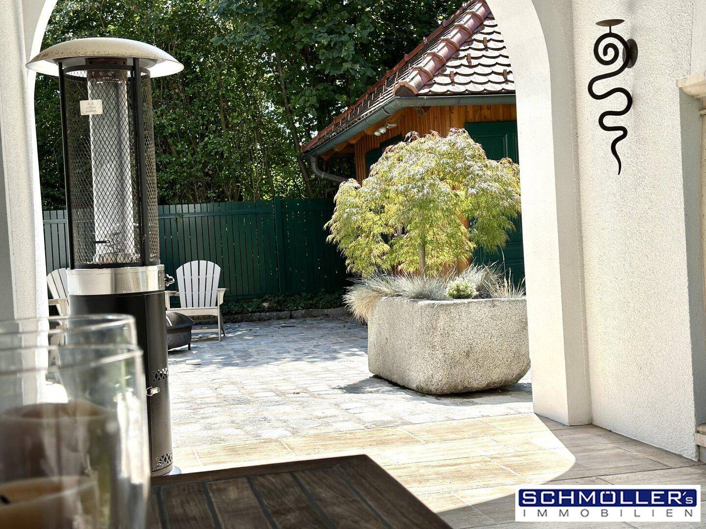 Sonniger Terrassenbereich mit Steinpflaster, großem Pflanzgefäß und Sitzgelegenheiten.
