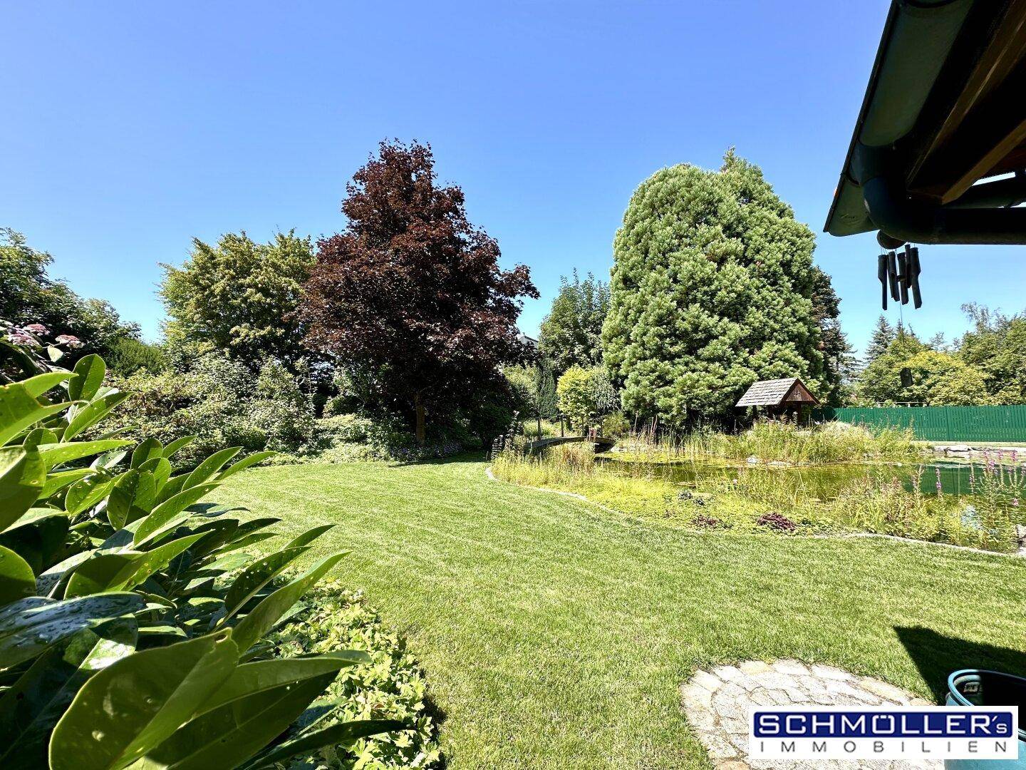 Weitläufiger Gartenbereich mit gepflegtem Rasen, einem Teich und hohen Bäumen unter blauem Himmel.
