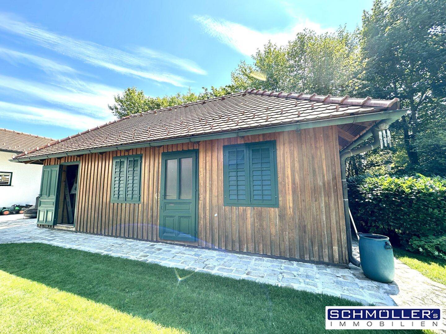 Rustikales Holzgebäude mit grünen Fensterläden und Ziegeldach im Gartenbereich.