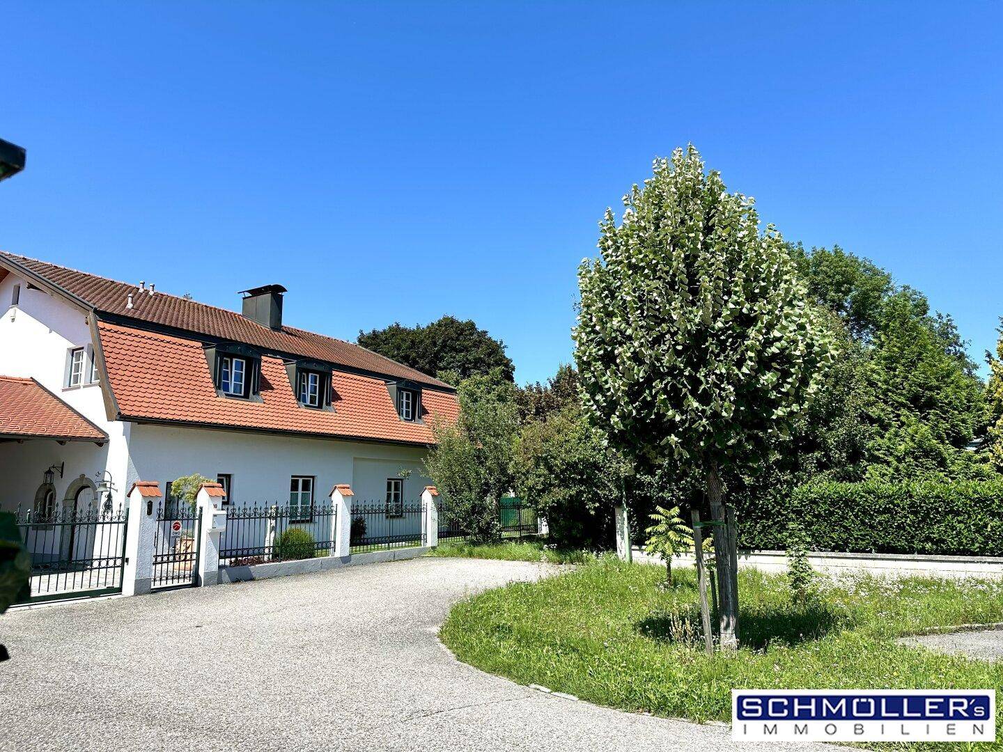 Vorderansicht des Hauses mit Zufahrt, Zaun und gepflegter Grünfläche unter strahlend blauem Himmel.