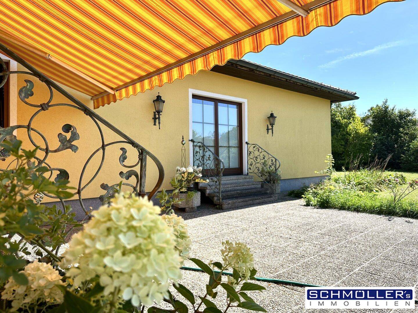 Sonniger Terrassenbereich mit gestreifter Markise, schmiedeeisernem Geländer und gepflastertem Boden.