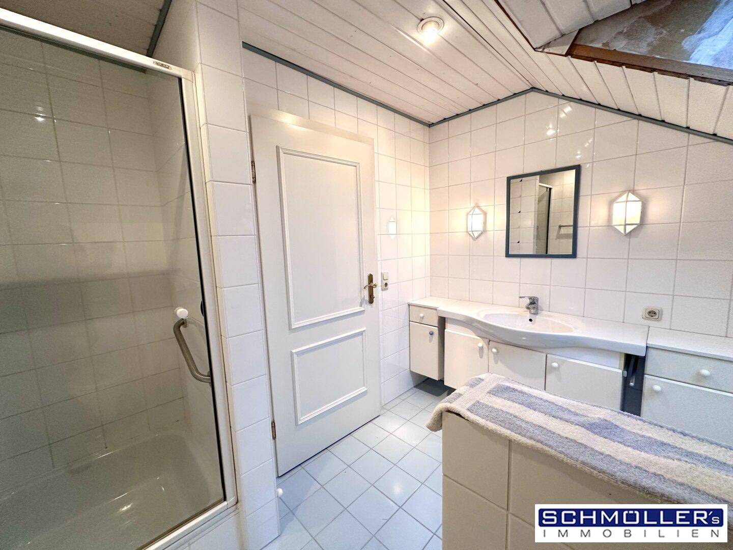 Badezimmer mit Duschkabine, Waschtisch und hellen Fliesen unter einer schrägen Dachfläche.
