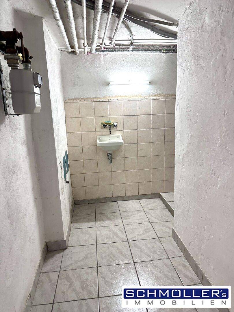 Kellerraum mit Fliesenboden, Waschbecken und sichtbaren Rohren an der Decke.