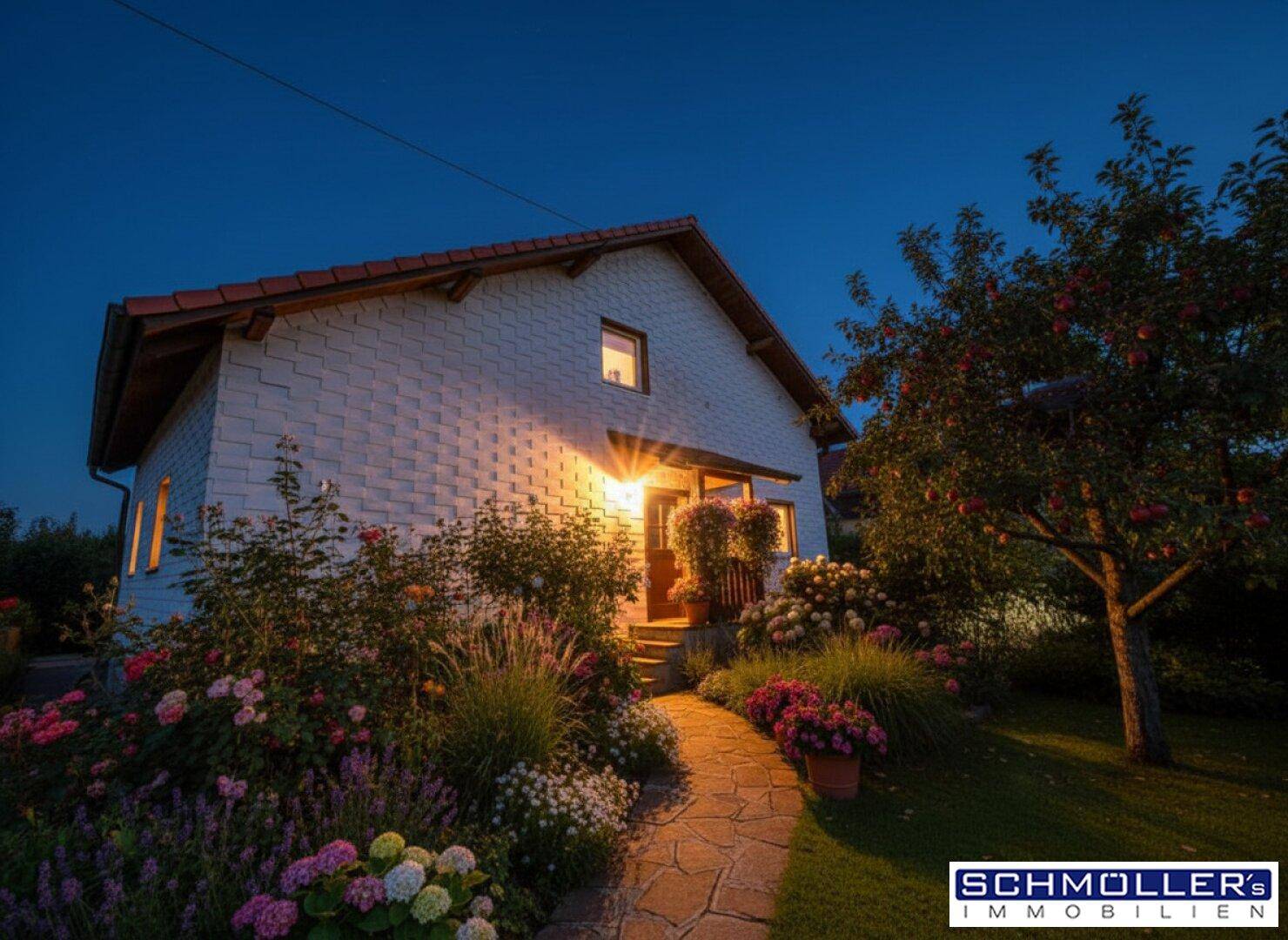 Haus bei Nacht mit beleuchtetem Eingangsbereich und einem blühenden Gartenweg.