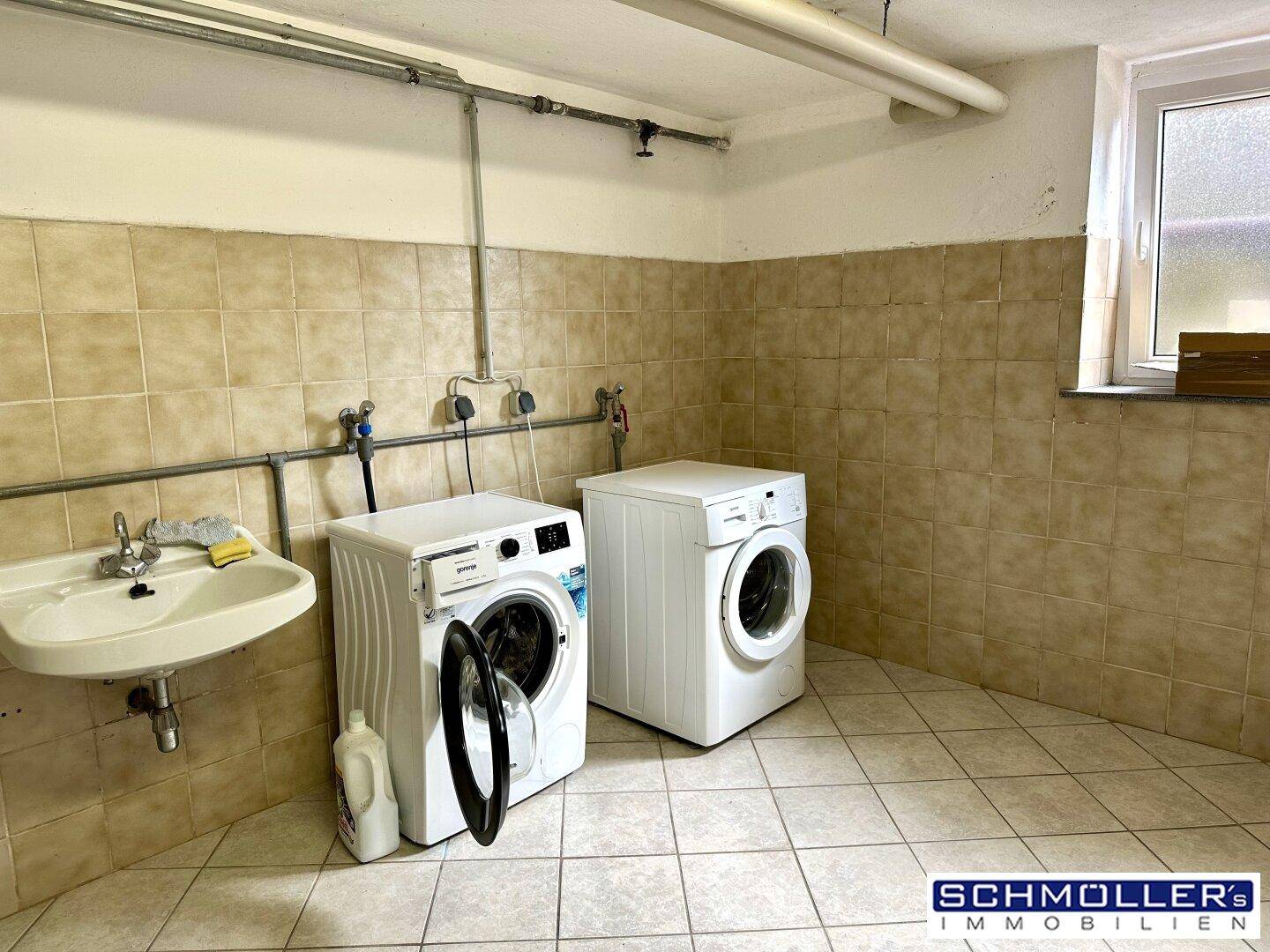 Waschküche im Keller mit Waschmaschinen, Waschbecken und Fliesenboden.