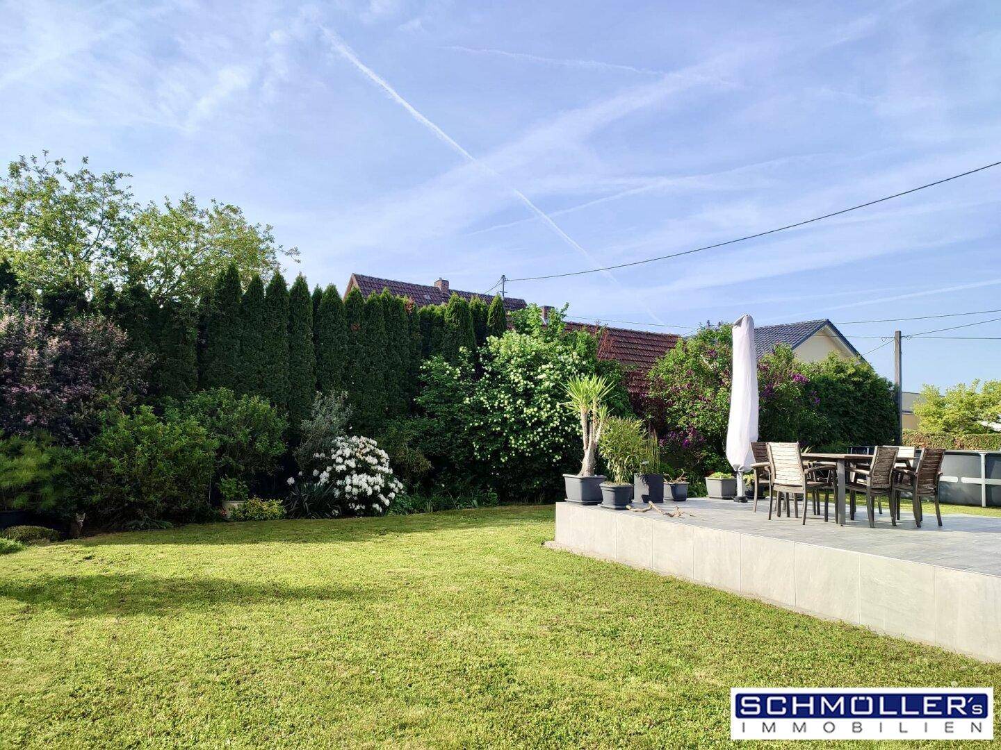 Sonniger Garten mit gepflegtem Rasen, hoher Hecke und einem Teil der Außenterrasse.