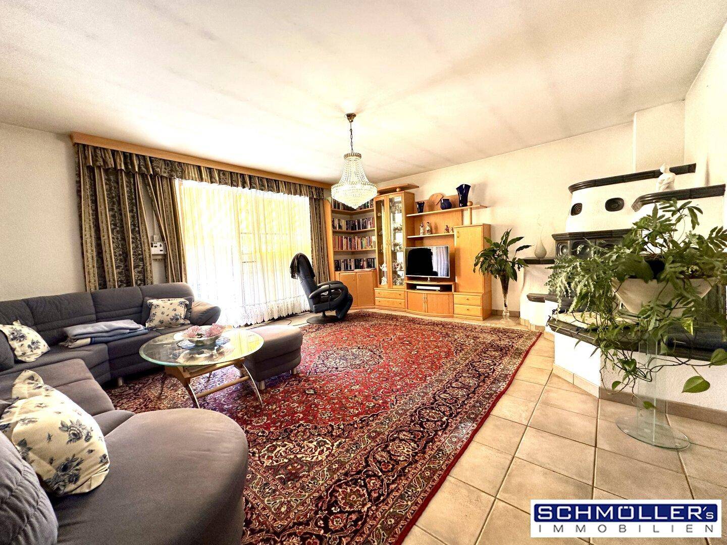 Geräumiges Wohnzimmer mit gemütlicher Sitzecke, traditionellem Kachelofen und viel Tageslicht durch große Fenster.