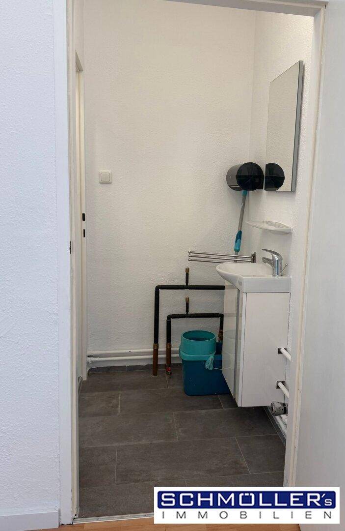 Kleiner Waschraum mit Waschbecken, Spiegel und sichtbaren Rohrleitungen auf dunklem Fliesenboden.