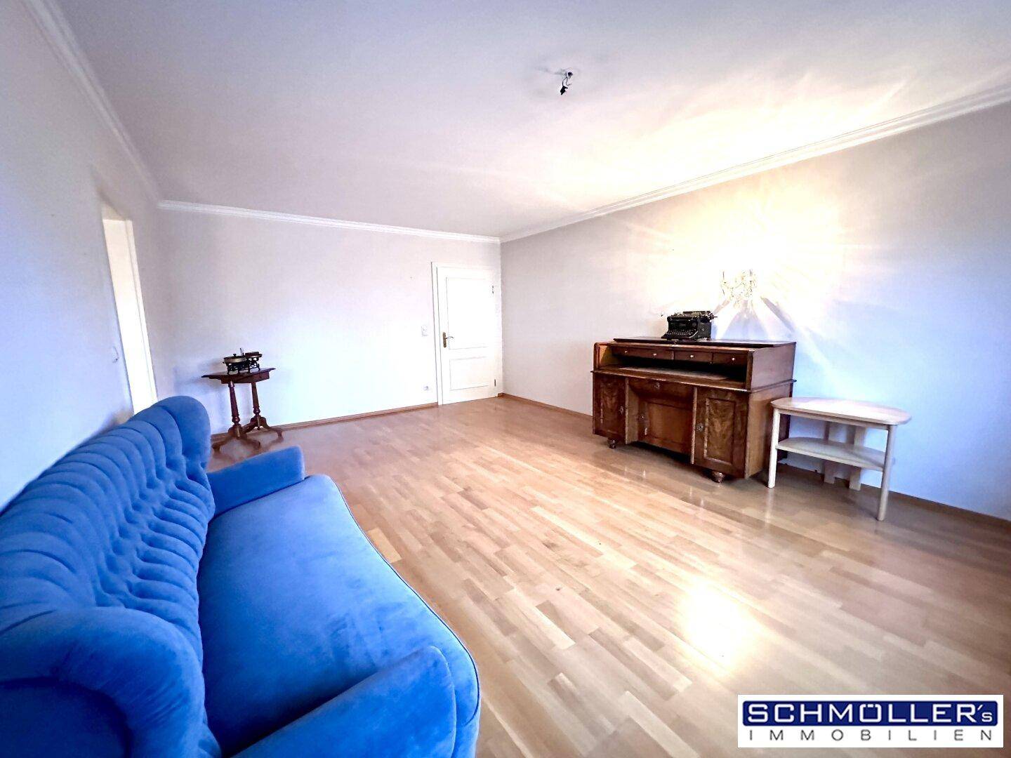 Wohnzimmer mit blauem Sofa, Parkettboden und einem antiken Schreibtisch unter hellem Licht.