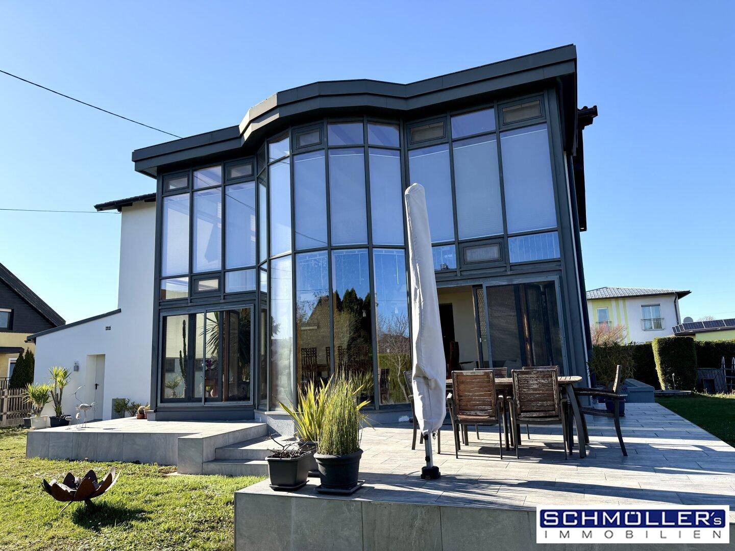 Geräumige Terrasse mit Gartenmöbeln und großem Sonnenschirm vor dem verglasten Anbau des Hauses.