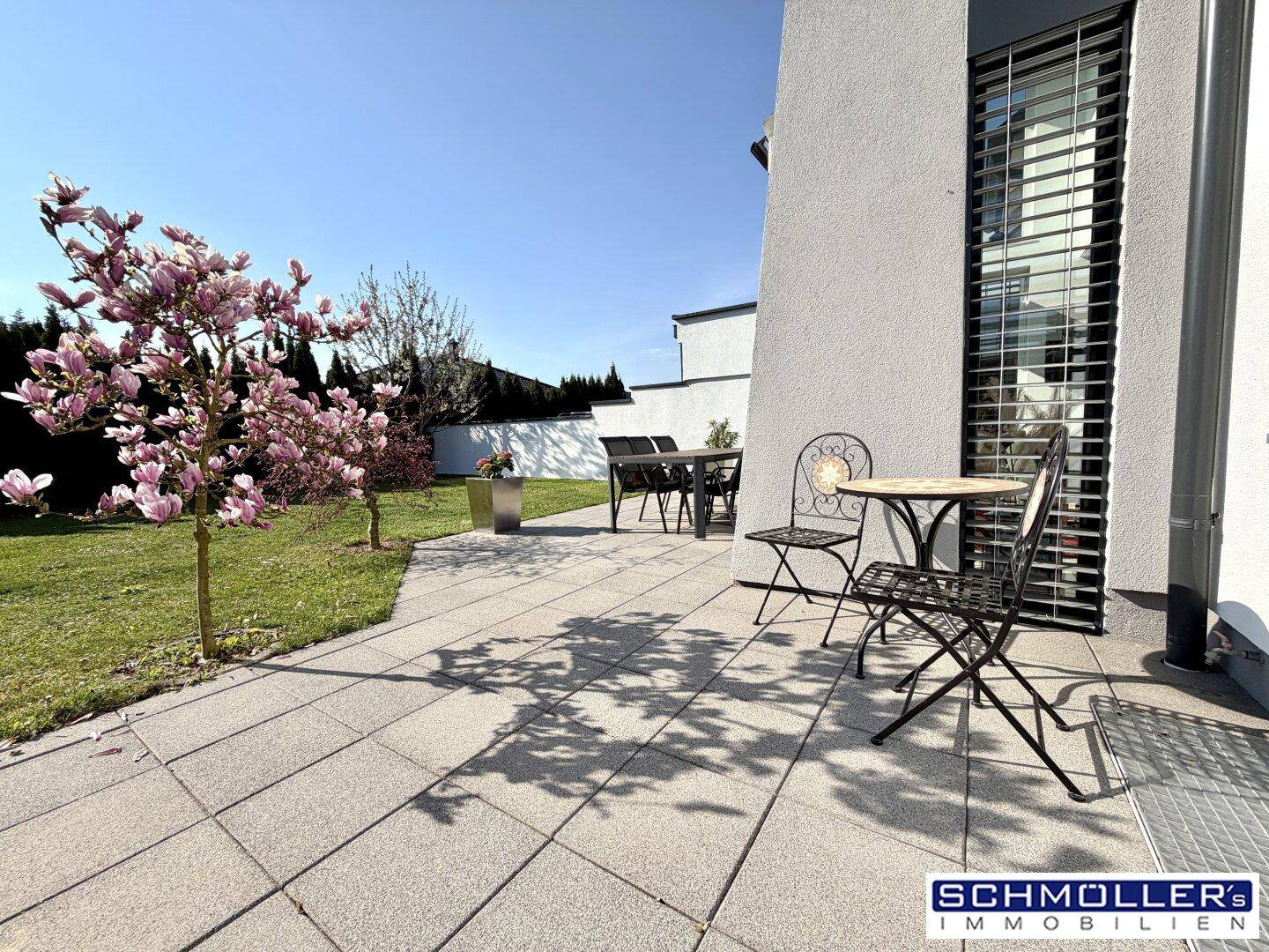 Detailansicht der gepflasterten Terrasse mit Gartenmöbeln und einem blühenden Magnolienbaum.
