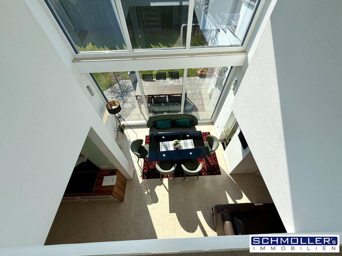 Blick von oben auf den lichtdurchfluteten Essbereich mit großen Fensterfronten und hohen Decken.