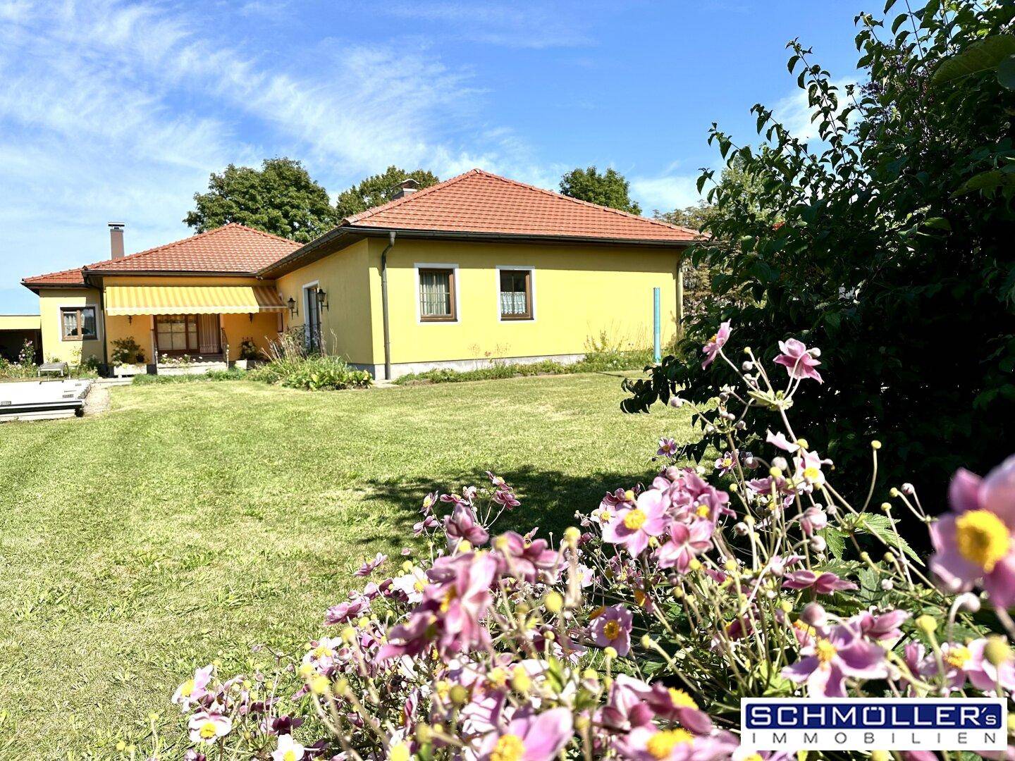 Gelbes Einfamilienhaus mit rotem Ziegeldach, umgeben von einem gepflegten Garten mit Blumen und einer überdachten Terrasse.