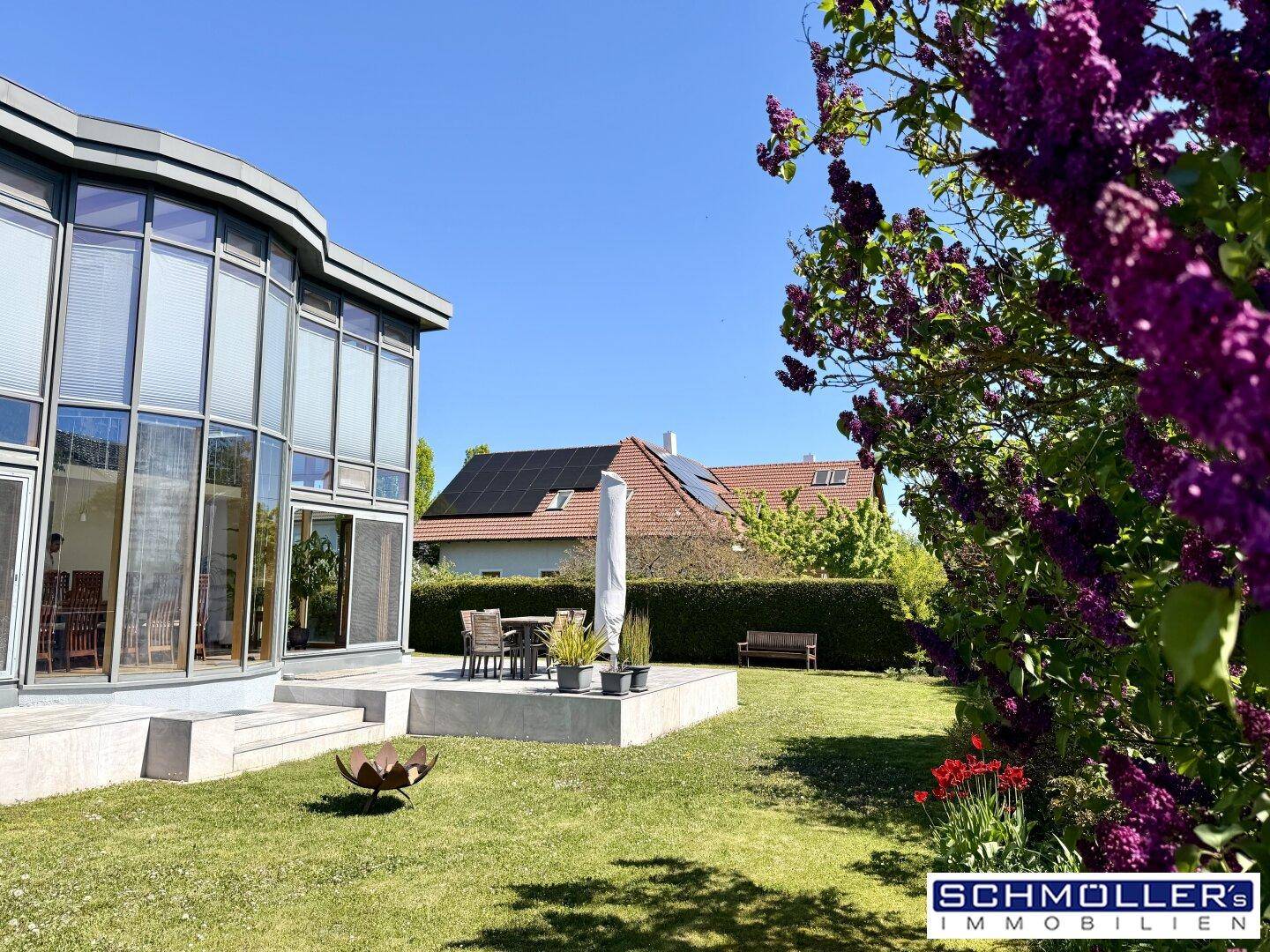 Moderner Anbau mit Glasfassade, möblierter Terrasse und gepflegtem Garten mit blühenden Pflanzen.