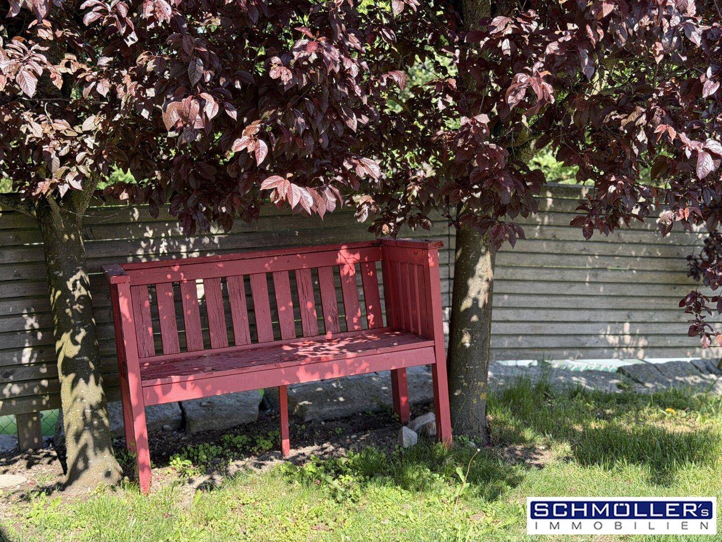 Rote Gartenbank unter einem Baum mit roten Blättern in einem sonnigen Gartenbereich.