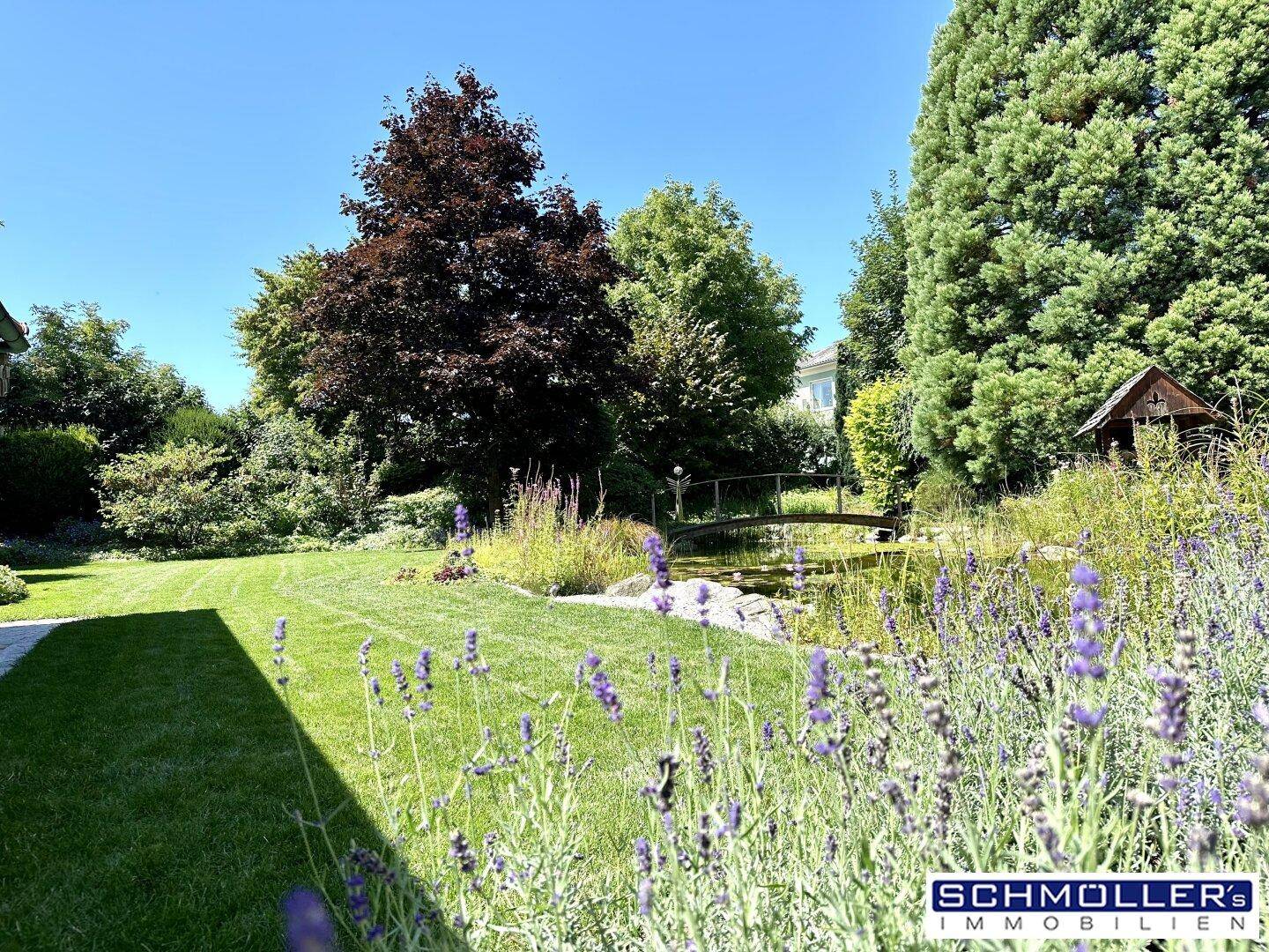 Gepflegter Gartenbereich mit blühendem Lavendel, Rasenfläche und einem kleinen Teich mit Brücke.