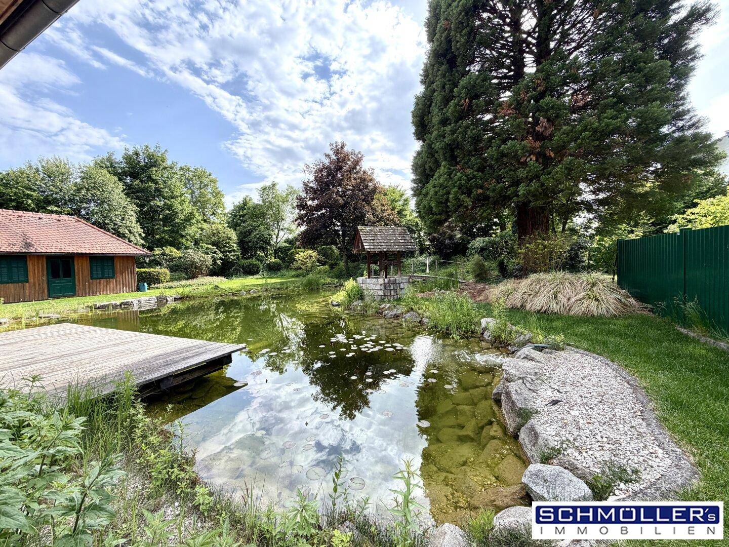 Idyllischer Teich mit Seerosen und Holzsteg, umgeben von üppigem Grün und einem Gartenhaus.