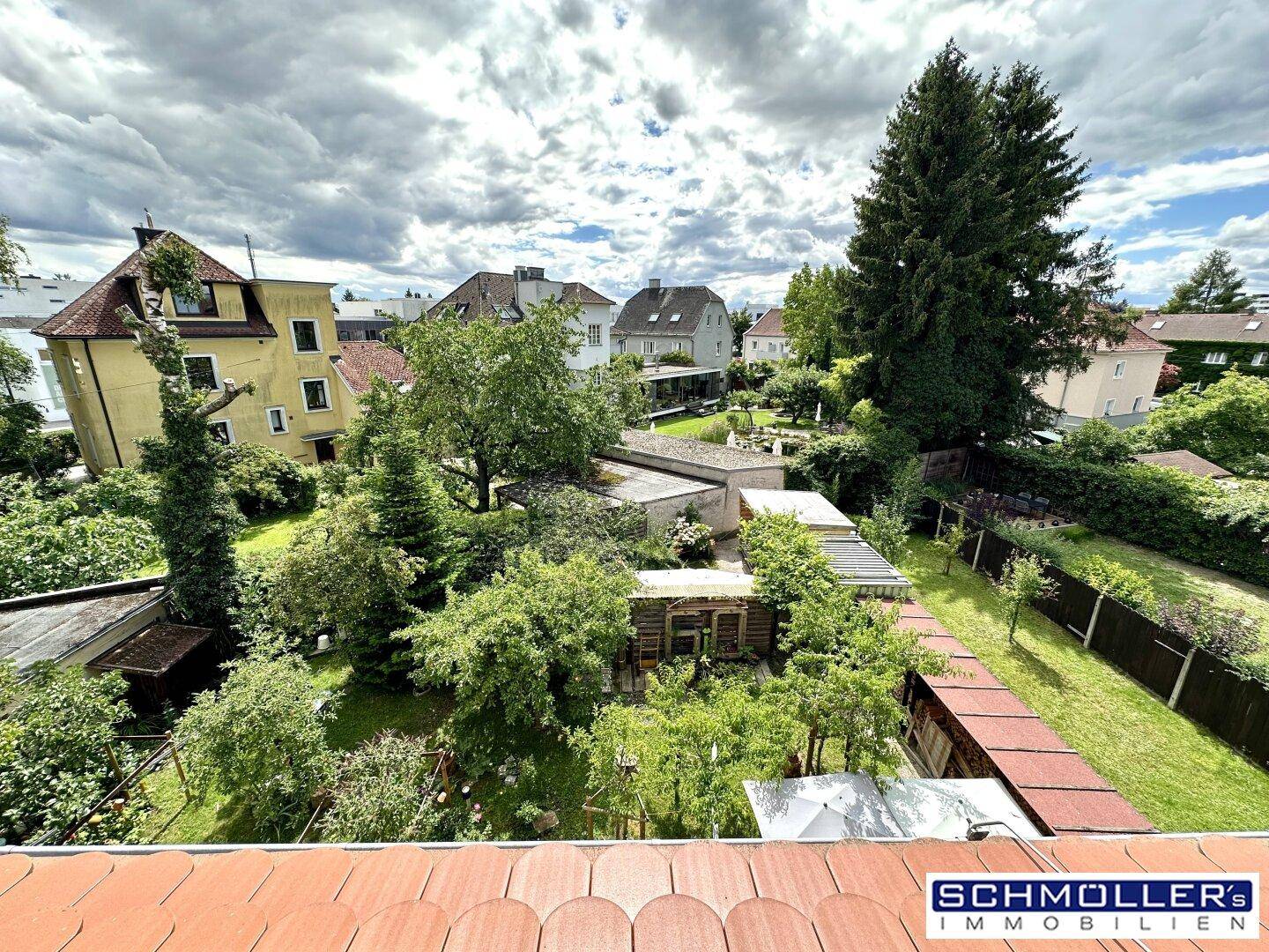Weitläufiger Blick über grüne Gärten und umliegende Häuser unter bewölktem Himmel.