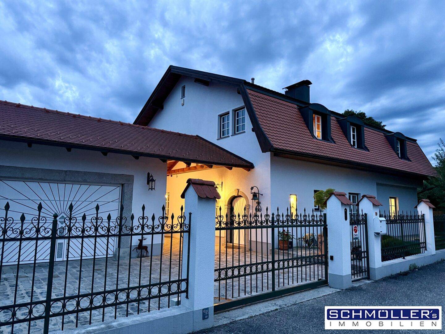 Elegantes Landhaus mit schmiedeeisernem Tor, Doppelgarage und beleuchtetem Eingangsbereich am Abend.
