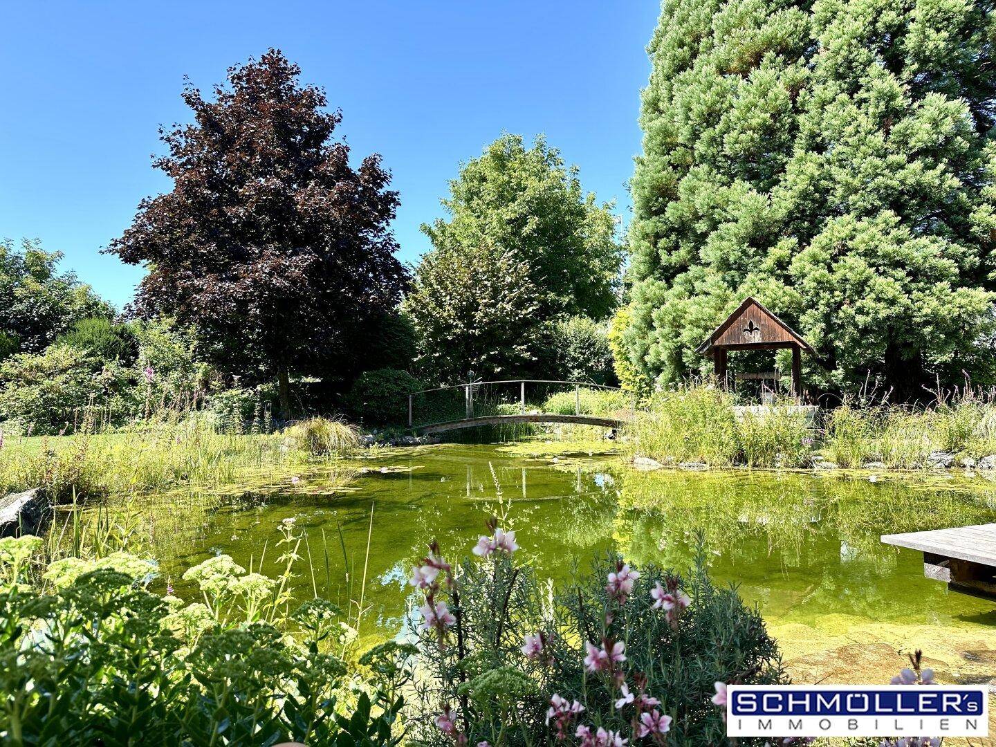 Idyllischer Garten mit großem Teich, kleiner Brücke und üppiger Vegetation unter blauem Himmel.