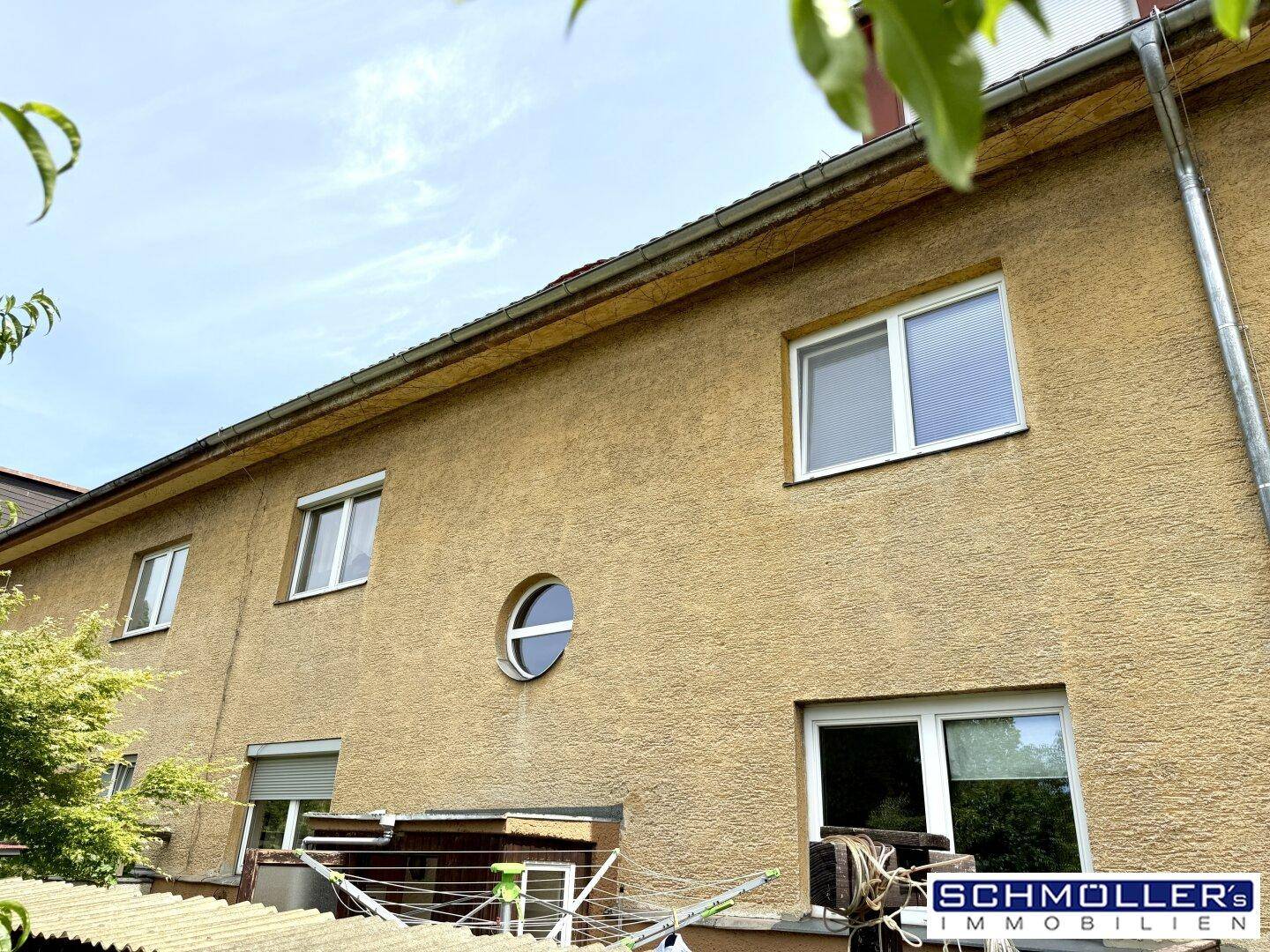 Rückseite eines Reihenhauses mit runden und rechteckigen Fenstern sowie einer Wäscheleine im Garten.