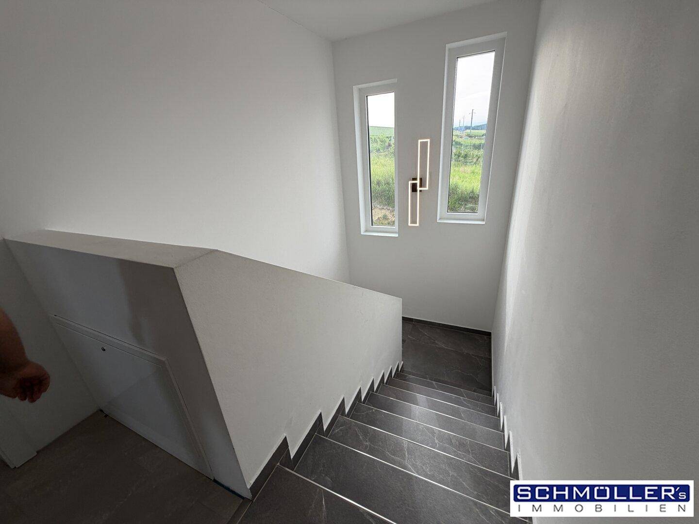 Moderner Treppenaufgang mit dunklen Stufen, weißen Wänden und hohen Fenstern.