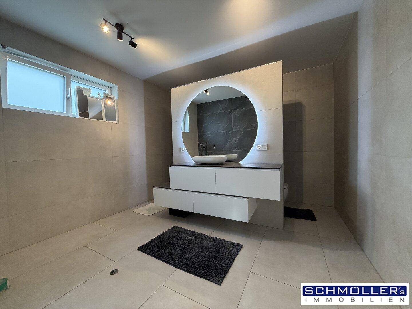 Modernes Badezimmer mit Doppelwaschtisch, beleuchtetem Spiegel und großen Fliesen.