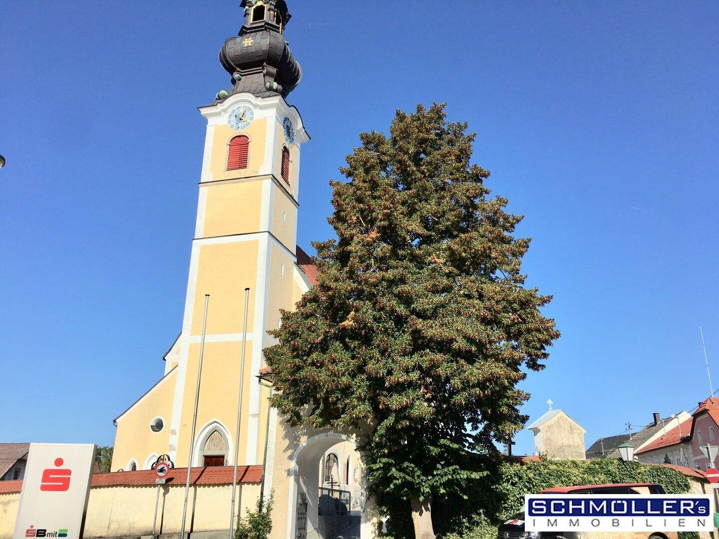 Historische Kirche mit markantem Turm und Uhr, umgeben von einem großen Baum.
