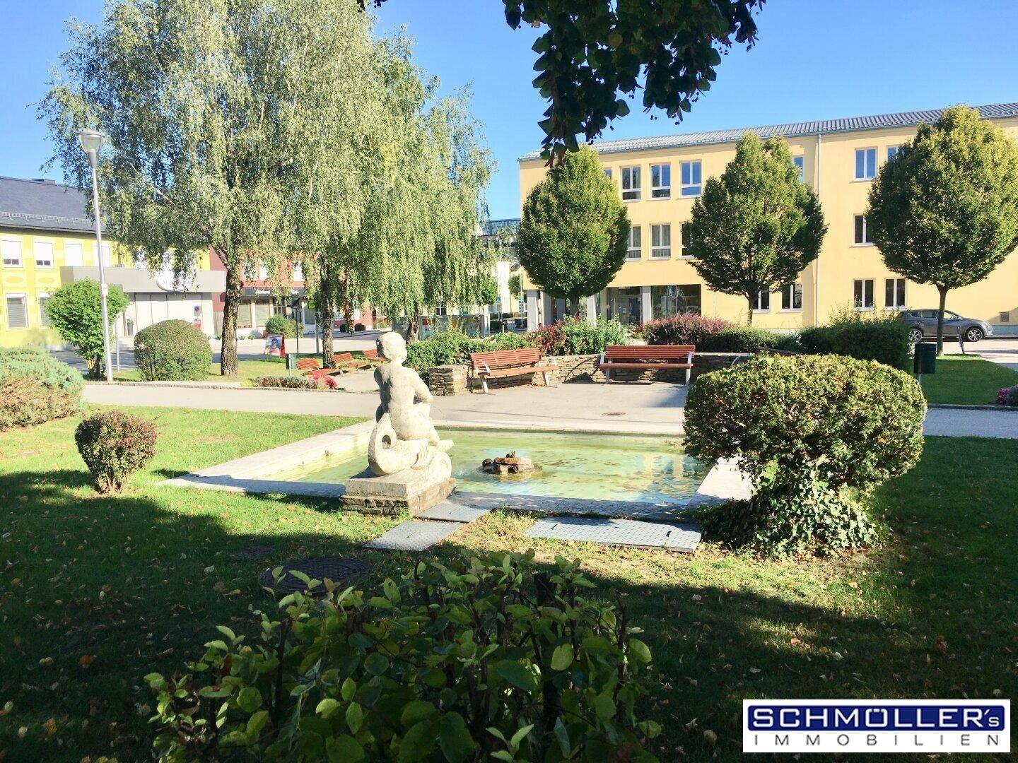 Öffentlicher Park mit einem Brunnen, Sitzbänken und schattenspendenden Bäumen.
