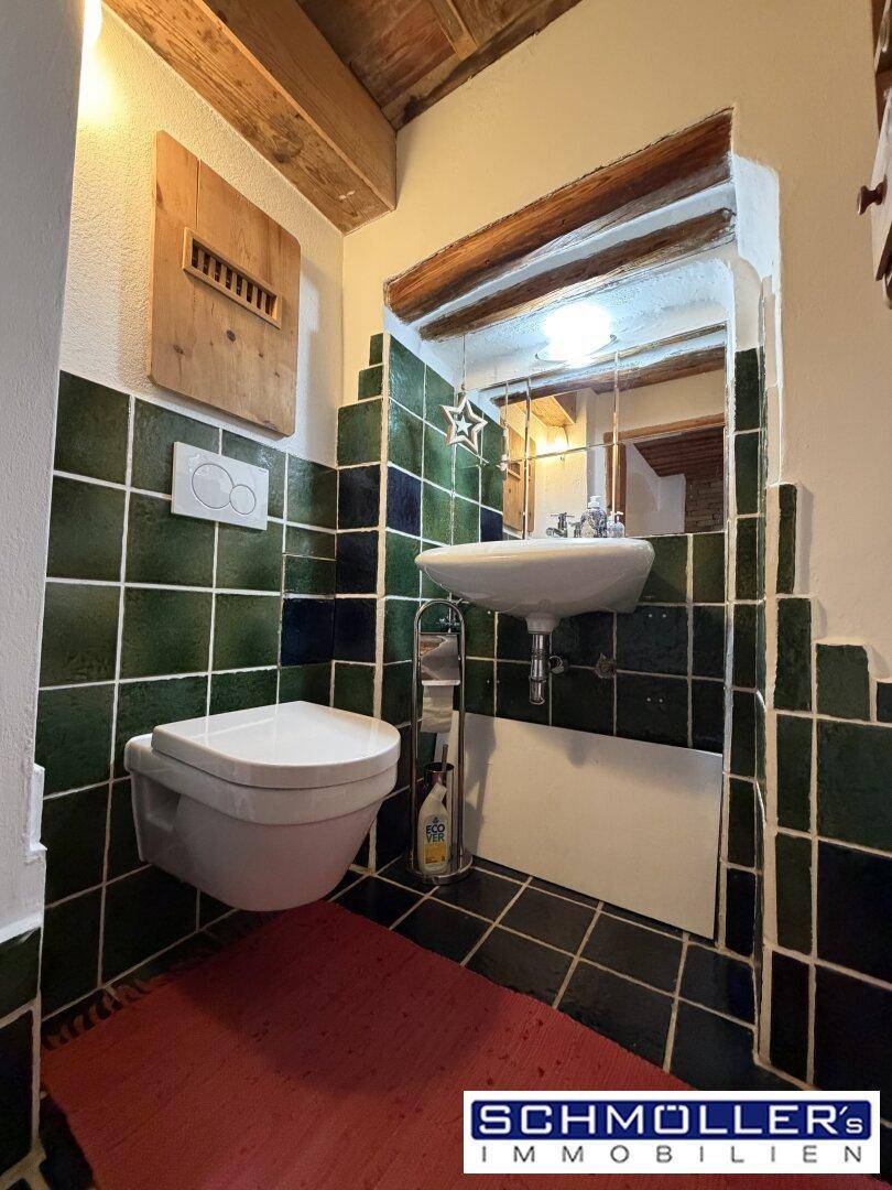 Kleines WC mit grünen Fliesen, wandhängender Toilette, kleinem Waschbecken und Holzbalkendecke.