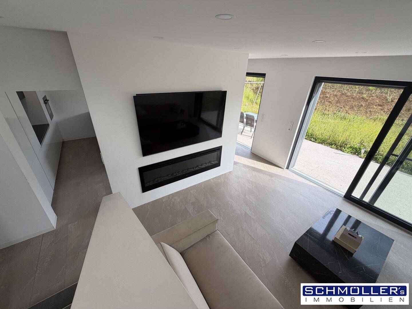 Helles Wohnzimmer mit modernen Fliesen, großem Fernseher und Zugang zum Gartenbereich.