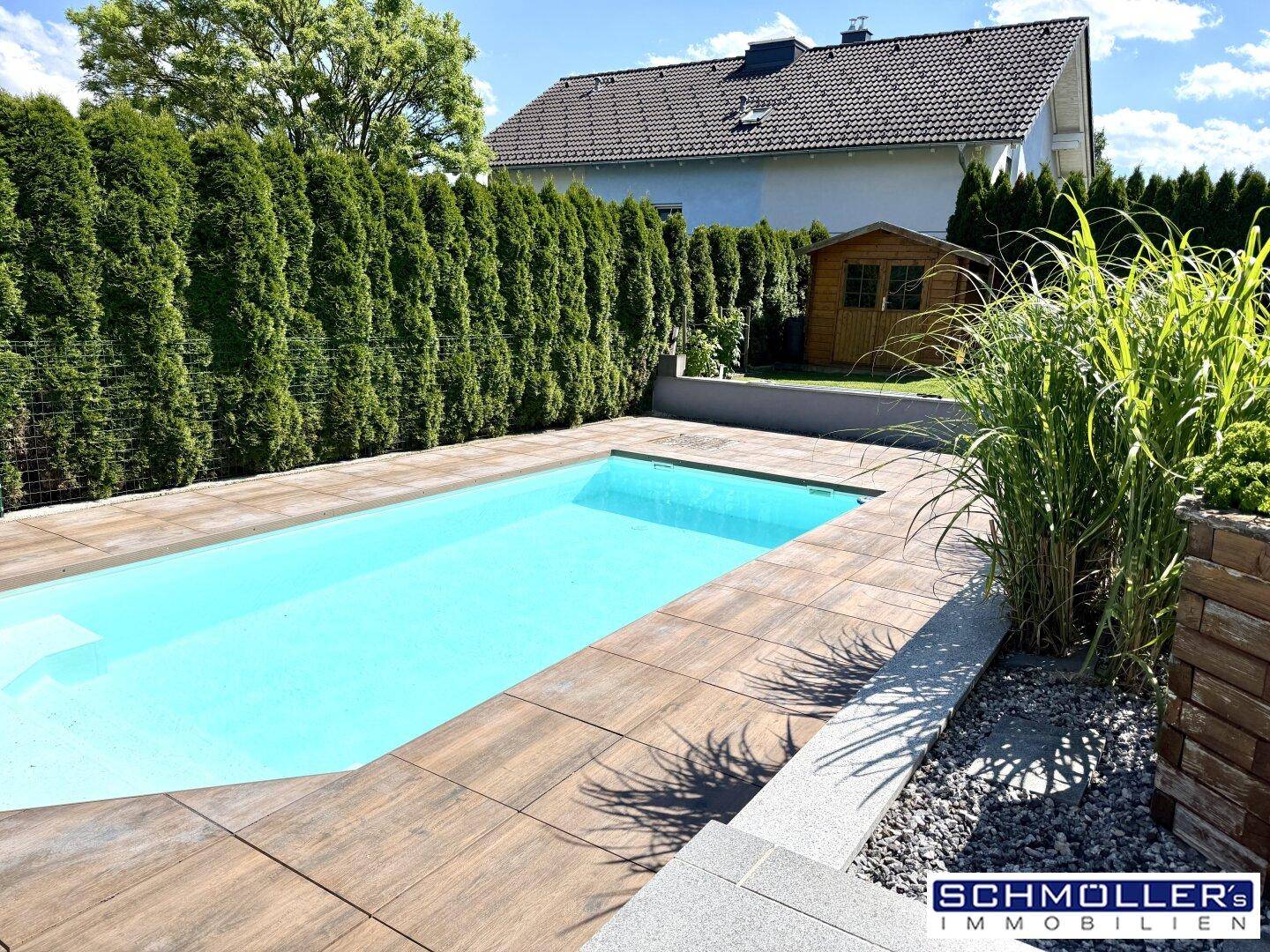 Außenpool mit gefliester Terrasse, umgeben von einer hohen Hecke und einem Gartenhaus.