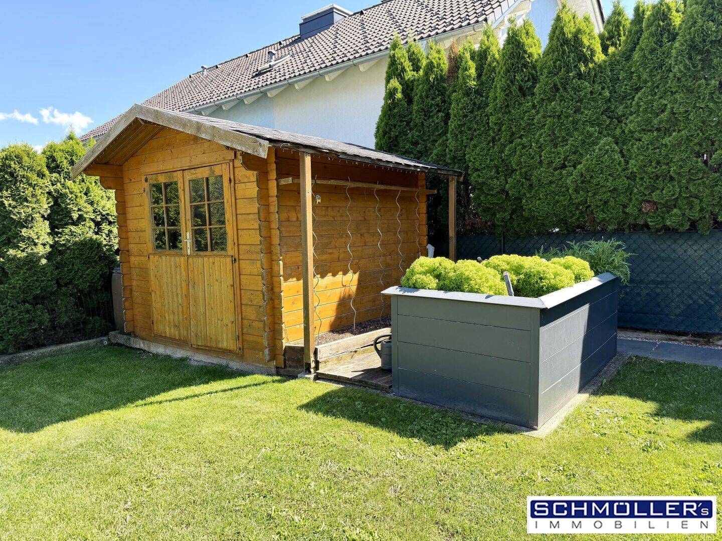 Kleiner Holzschuppen im Garten neben einem Hochbeet, umgeben von grüner Hecke.