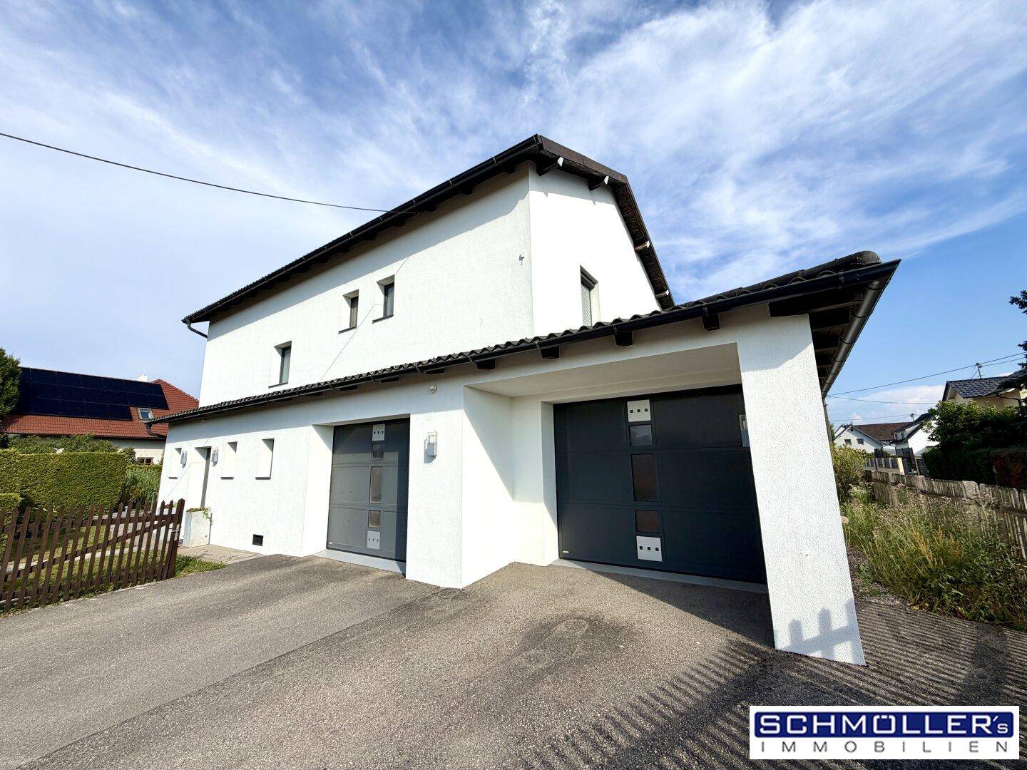 Modernes weißes Einfamilienhaus mit zwei Garagentoren und Satteldach unter blauem Himmel.