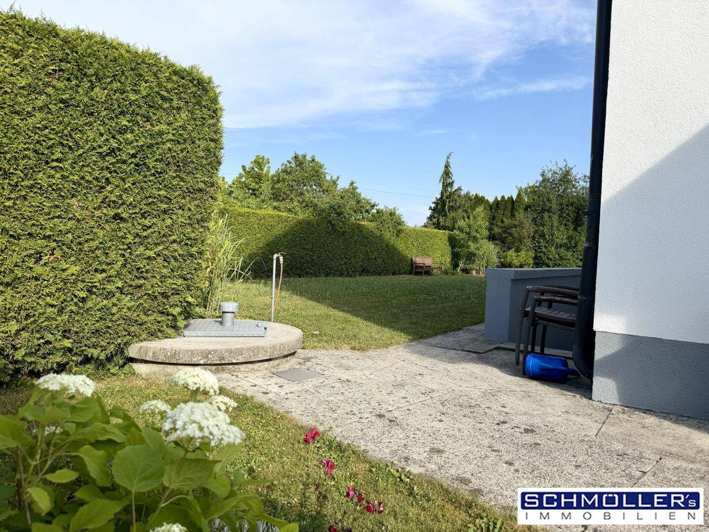 Gepflegter Gartenbereich mit grüner Rasenfläche, hoher Hecke und blühenden Pflanzen unter blauem Himmel.