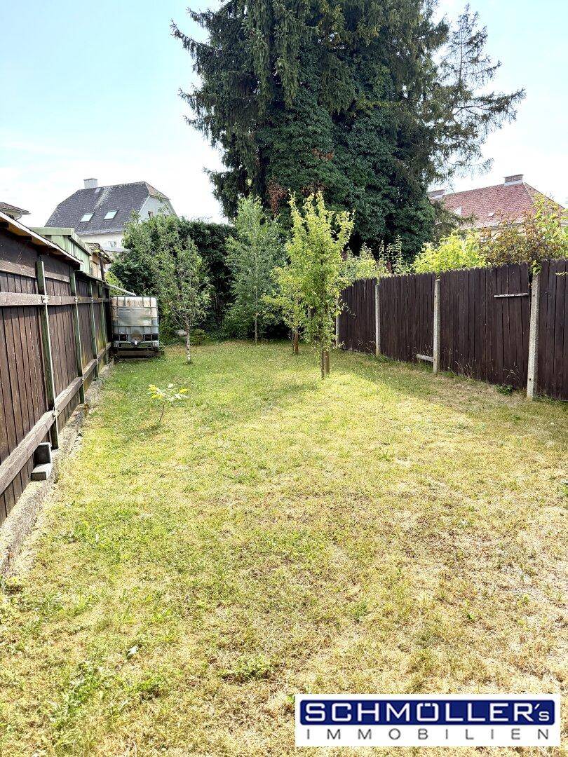 Gepflegter Gartenbereich mit Rasenfläche und hohen Bäumen, umgeben von Holzzäunen.