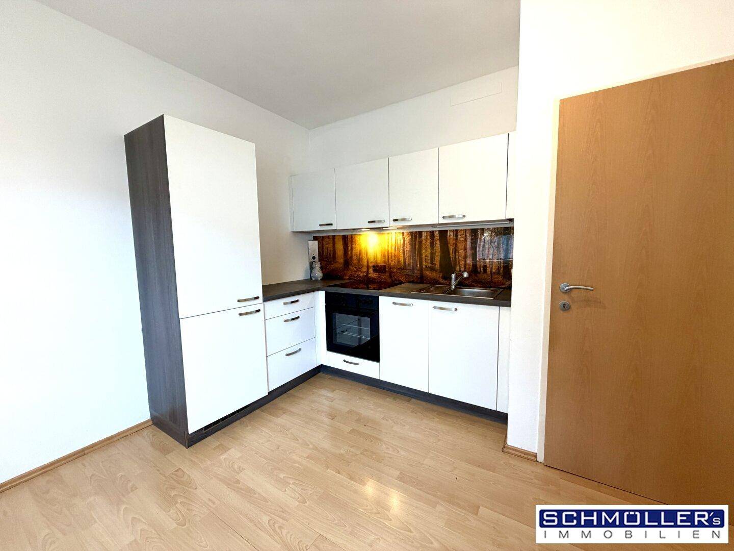 Kompakte Küche mit weißen Schränken, integriertem Backofen und dekorativer Wald-Rückwand.
