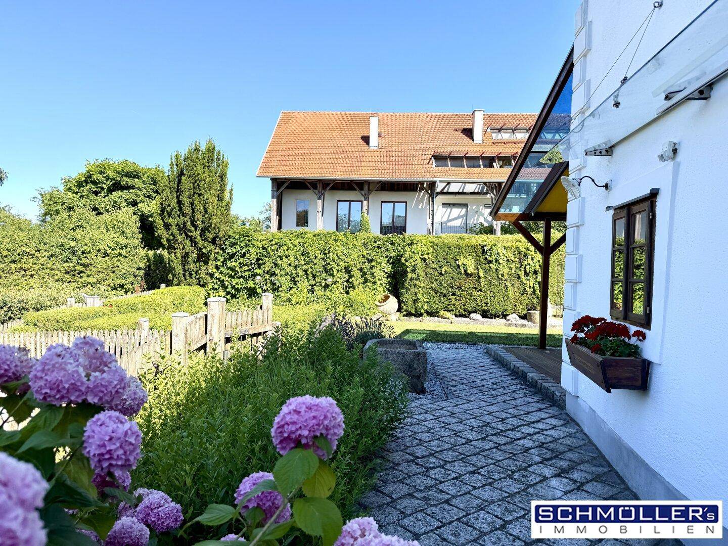 Gepflasterter Gartenweg führt entlang des Hauses mit blühenden Hortensien und Hecken.